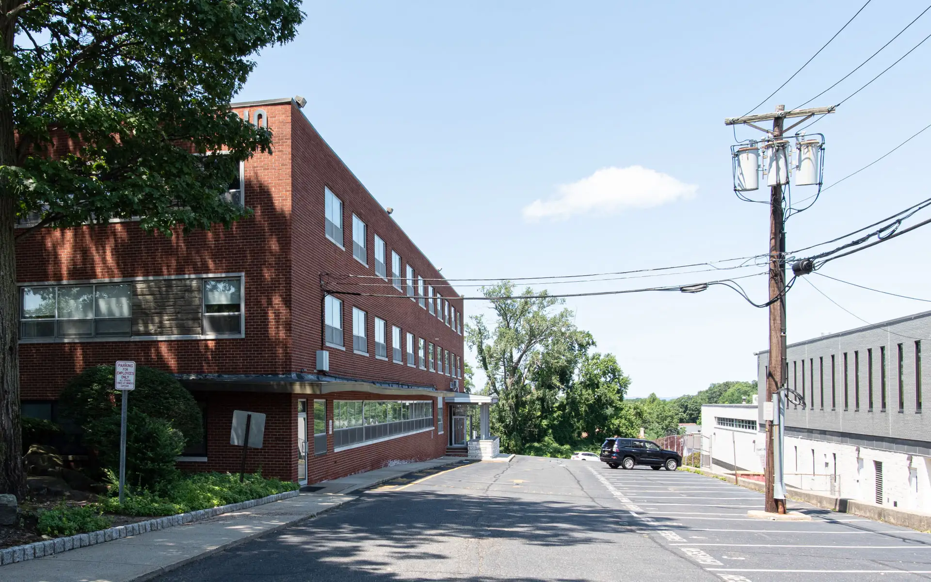 Exterior view of a red brick industrial building under clear daylight with parking spaces and surrounding trees.