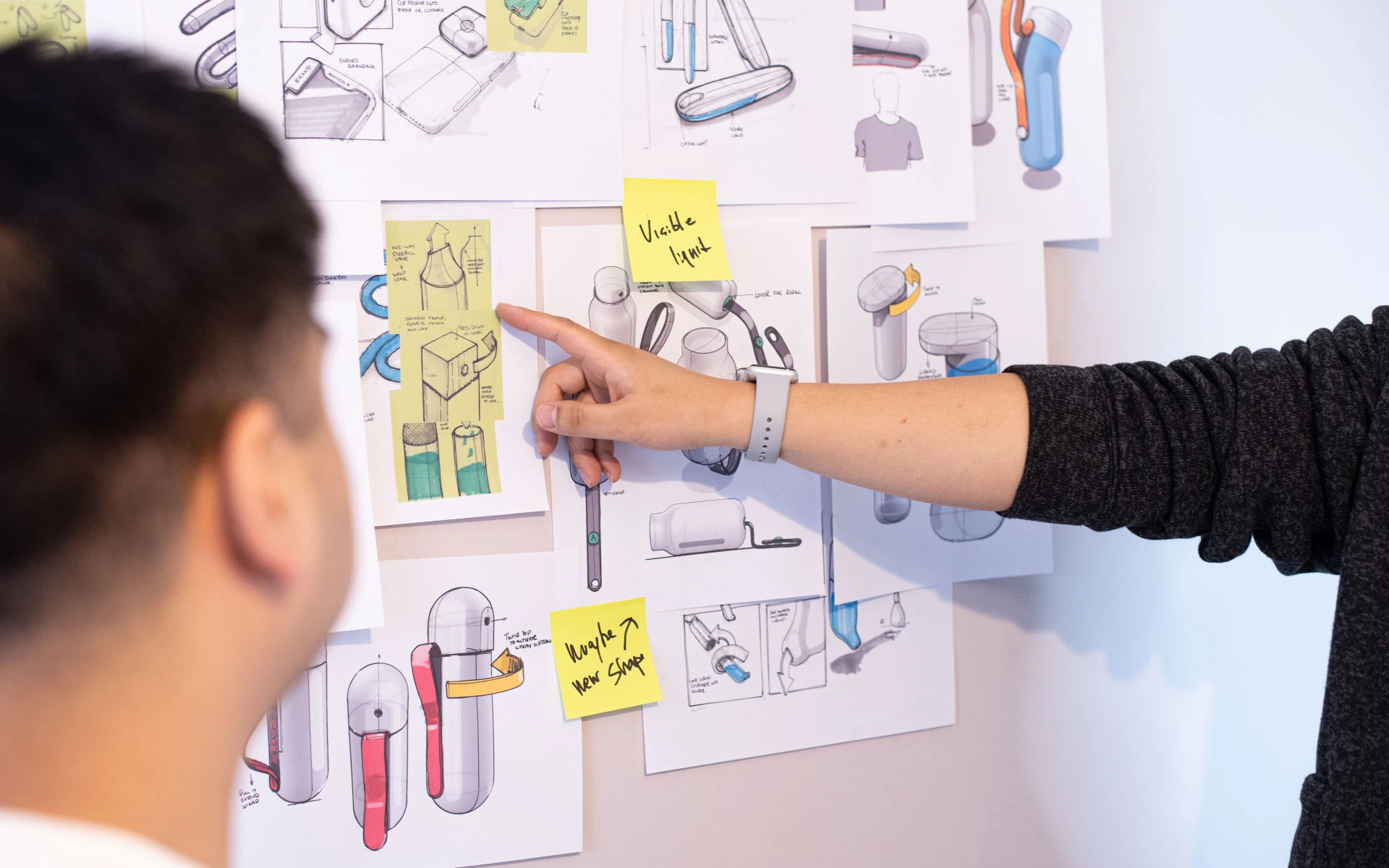 Designers reviewing product concept sketches on a wall covered with industrial design drawings and notes.