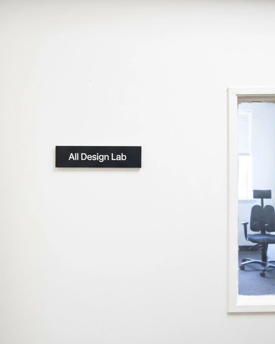 Minimal white office wall with an “All Design Lab” sign next to a window showing an empty office chair inside.