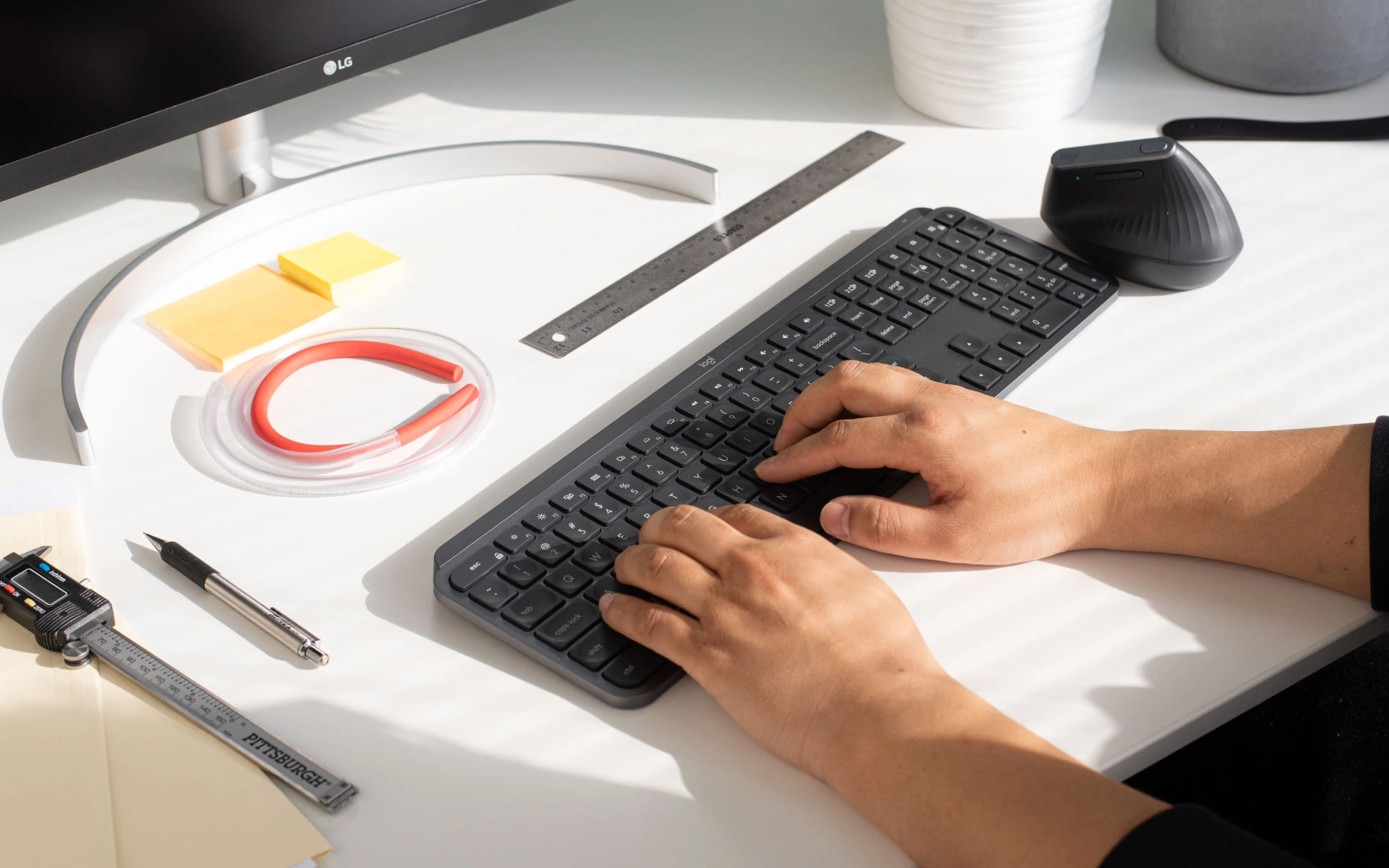 Designer typing at a clean workstation with a keyboard, ruler, caliper, and orange prototype component under natural light.