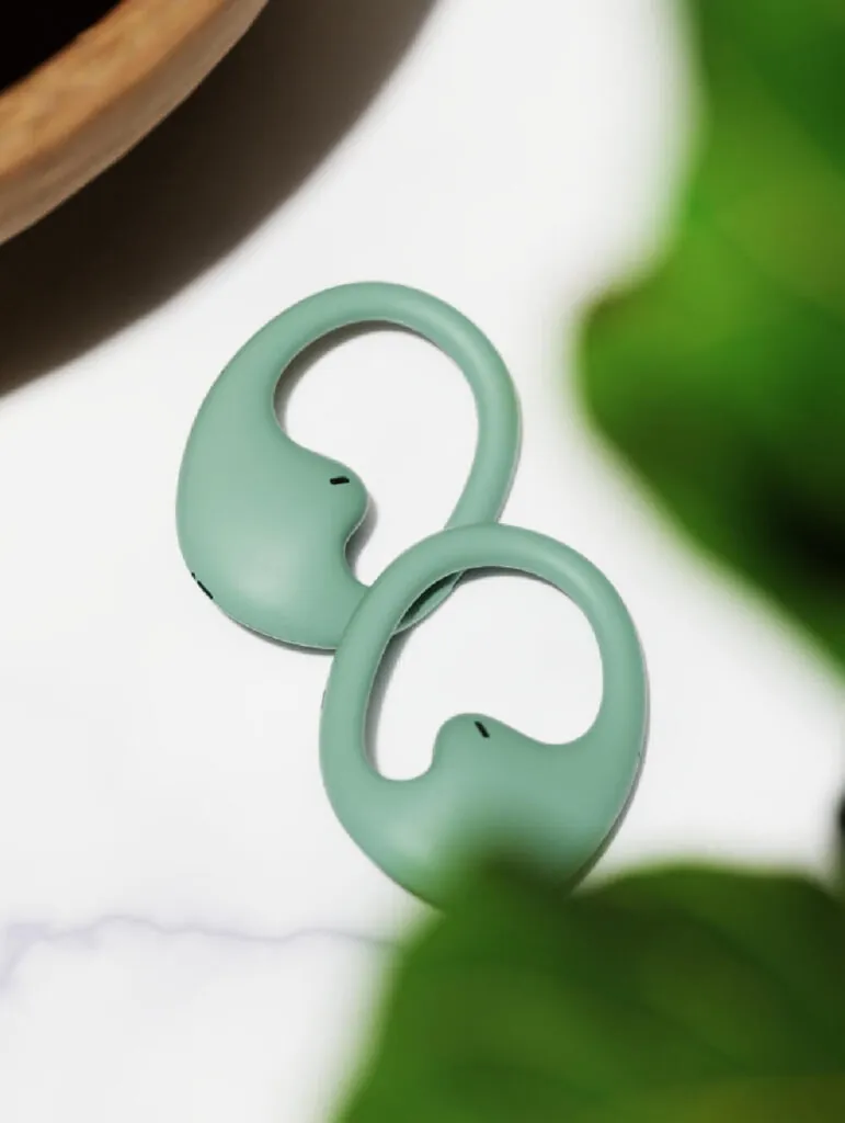 Two green ring like objects with a sleek design, amidst soft green leaves. Earbugs Headphones by All Design Lab