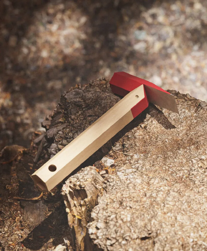 A hammer with a red top and a wooden handle on a tree stump. Hex tools hammer by All Design Lab.