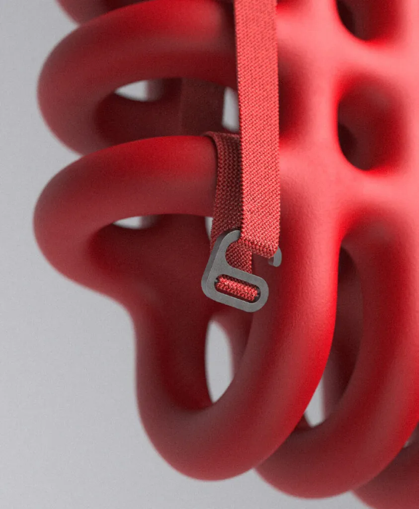 Close up of red foam cage like object and strap and buckle hanging off of it. Mega Mesh Backpack by All Design Lab.