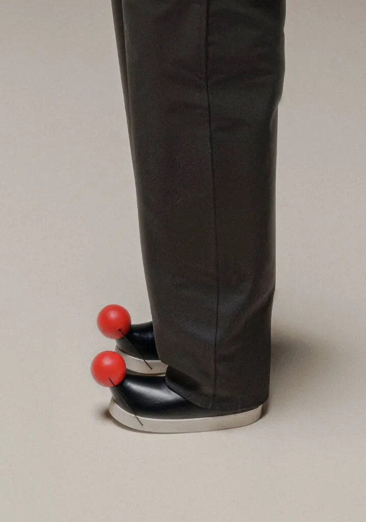 Red ball on black boots worn by person wearing grey pants. Pom footwear by All Design Lab.