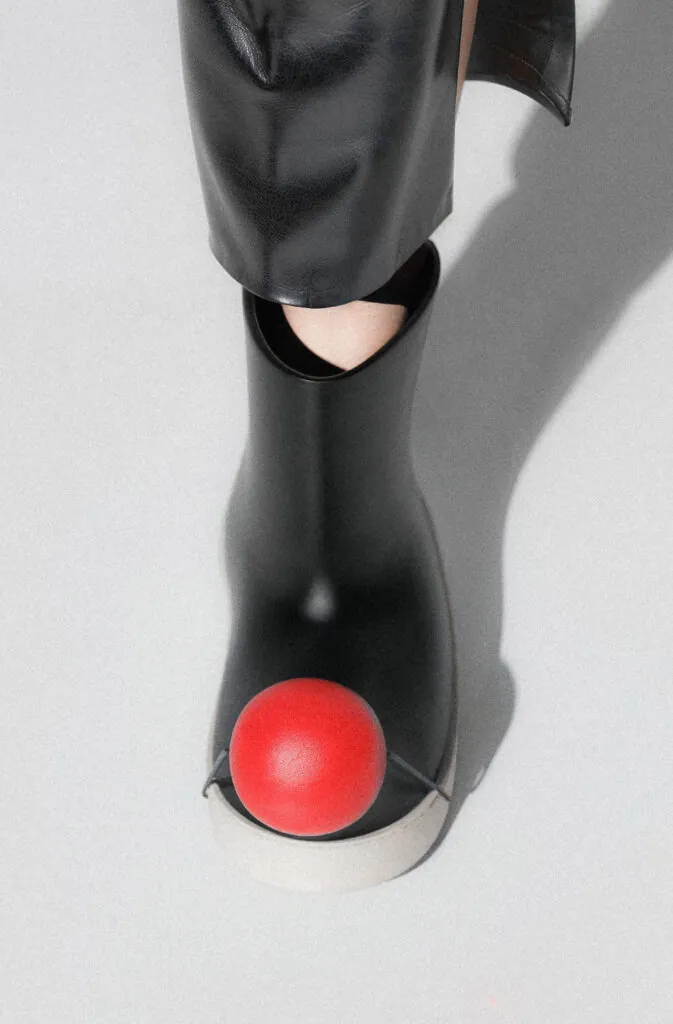Black boots with red ball on foot, white background. Pom footwear by All Design Lab.