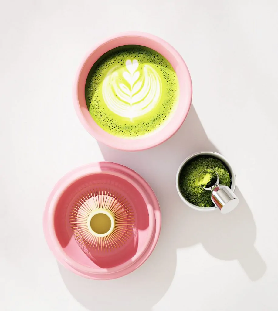Pink ceramic matcha bowl filled with bright green matcha latte, shown with whisk holder and scoop.