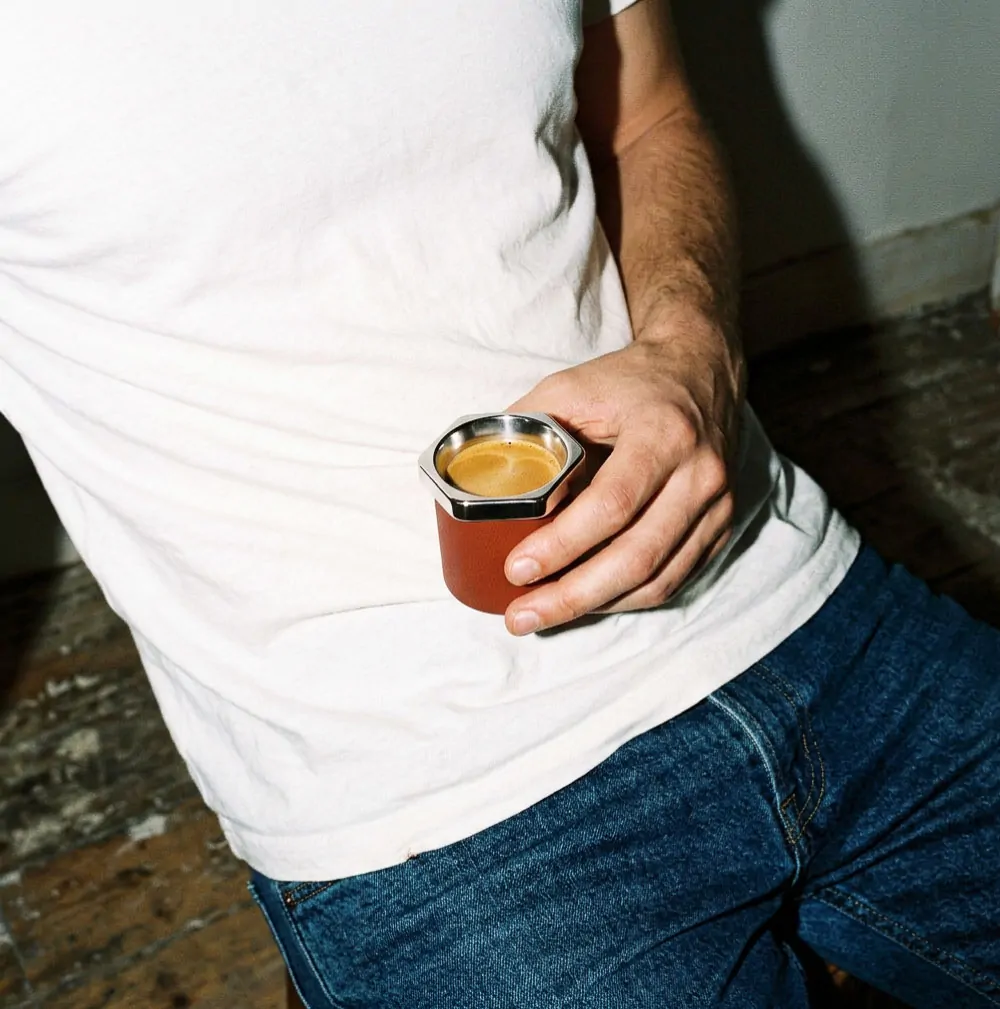 Hand holding Esagono espresso cup filled with espresso, burgundy powder-coated body against white t-shirt and denim jeans, shot on 35mm film